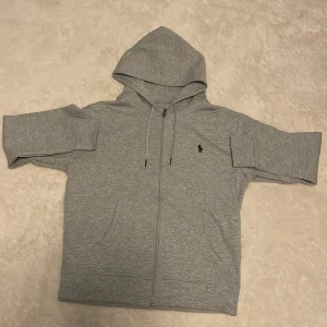 Zip tröjan från polo ralplauren - Snygg grå hoodie från Polo Ralph Lauren med dragkedja framtill, huva med snörning och klassisk logga på bröstet. Tröjan har korta ärmar och två fickor framtill. Perfekt för en chill och sportig look.
