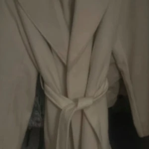 Beige kappa med bälte och krage - Stilren beige kappa med bred krage och knytskärp i midjan. Jackan har en klassisk, rak passform och är tillverkad i ett mjukt ull-liknande material. Perfekt för dig som vill ha en clean och tidlös look under höst och vinter.