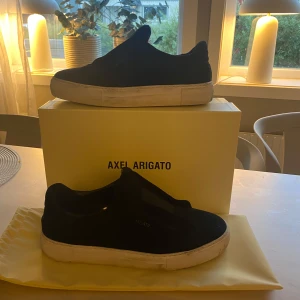 Svarta sneakers från Axel Arigato - Stilrena svarta sneakers från Axel Arigato med vit platt sula och diskret logga på sidan. Skorna har en minimalistisk design utan snörning och är tillverkade i mocka med elastiskt band över foten för en clean look.