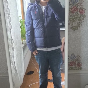Blå pufferjacka från Polo Ralph Lauren - Säljer en blå pufferjacka från Polo Ralph Lauren med huva och dragkedja. Jackan har en klassisk quiltad look, två fickor med dragkedja och den ikoniska röda loggan på bröstet. Perfekt för kyliga dagar och riktigt snygg till jeans.