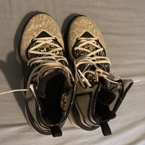 Nike LeBron 19 svart/vit sneakers - Säljer ett par Nike LeBron 19 sneakers i svart och vitt med coolt rutigt mönster och leoparddetaljer på plösen. Skorna har snörning, hög siluett och genomskinliga paneler på sidorna. Sulan är vit med synlig dämpning och LeBron-logga undertill. Perfekta för dig som gillar unika basketskor.