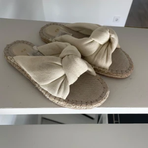 Beige sandaler med knut från H&M - Snygga sandaler från H&M i beige bomullstyg med breda band och stor knut upptill. Sulan är i grovt vävt material med synliga sömmar runt kanten. Perfekta för sommaren och ger en chill vibe till din outfit.
