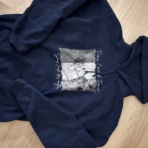 Mörkblå hoodie med tryck Paris - Mörkblå hoodie med huva och snörning. Framsidan har ett coolt tryck i grått, vitt och svart med texten 'Paris, Cœur de France' och grafiska detaljer. Perfekt för dig som gillar streetstyle och vill ha en bekväm tröja med snygg design.
