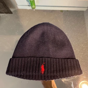 Mörkblå stickad mössa Polo Ralph Lauren - Mörkblå stickad mössa från Polo Ralph Lauren med klassisk röd broderad logga framtill. Mössan har uppvikt kant med ribbad struktur och är tillverkad i mjuk ull. Perfekt för kalla dagar och en clean streetstyle-look.