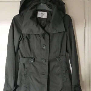 Svart trenchcoat med huva från ONLY - Stilren svart trenchcoat från ONLY med stor krage, huva och knappar framtill. Jackan har markerad midja med dekorativa knappar vid fickorna och långa ärmar. Perfekt för dig som gillar en klassisk look med modern twist.