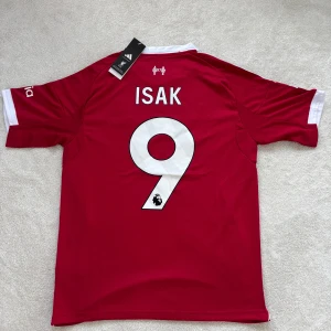 Liverpool Isak #9 Adidas fotbollströja - Liverpool FC matchtröja från Adidas med Isak och nummer 9 på ryggen. Röd med vita detaljer, broderad Adidas-logga, Premier League-märke i guld på ärmen och Standard Chartered-tryck på bröstet. Tillverkad i ventilerande polyester för maximal komfort.