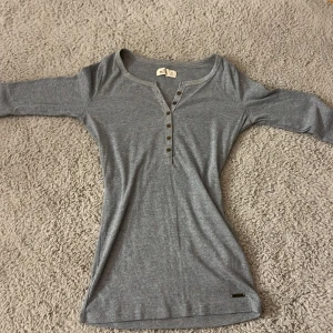 Grå ribbad topp från Hollister XS - Grå ribbad långärmad topp från Hollister med snygga knappar framtill och broderade blommor på ärmsluten. Mjuk och stretchig bomullsblandning, perfekt för lager-på-lager. Snygg passform och unika detaljer som sticker ut.