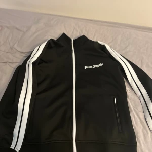 Palm Angels svart track jacket - Svart track jacket från Palm Angels med vita ränder längs ärmarna och vit dragkedja. Jackan har hög krage, broderad logga på bröstet och fickor med dragkedja. Sportig och clean look, perfekt för streetwear-stil.  Palm angels påse finns med, priset kan diskuteras 