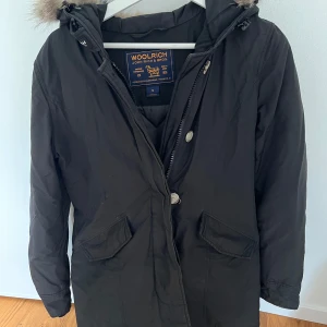 Svart parkajacka från Woolrich med huva - Svart parkajacka från Woolrich med klassisk huva.. Jackan har dragkedja och knappar framtill, två stora fickor med lock och är tillverkad i polyester och bomull. Perfekt för kalla dagar och riktigt snygg till vintern. Använd en säsong.