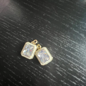 Guldiga örhängen med glittrande stenar - Säljer ett par snygga örhängen i guldton med fyrkantiga, glittrande stenar i mitten. Stenarna är klara och ger en lyxig vibe, perfekt för att lyfta din look. Designen är stilren och modern med en rektangulär form och bling-effekt. Man får endast det som är på bilden och en påse till och lite skydd till och lite mer men det är frakt.