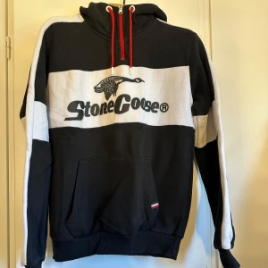 Svart och vit hoodie från Stone Goose - Snygg hoodie från Stone Goose i svart och vitt med breda blockränder och stor logga framtill. Tröjan har huva med röda snören, känguruficka och ribbade muddar. Perfekt för dig som gillar streetwear och vill sticka ut lite extra.