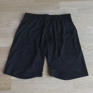 Svarta träningsshorts från Energetics - Svarta träningsshorts från Energetics i lätt och smidigt material. De har elastisk midja och är tillverkade i 100% polyester, perfekta för sport eller gym. Klassisk och enkel design utan mönster eller extra detaljer.