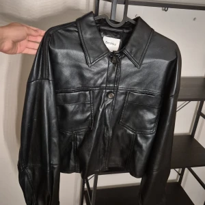 Svart skinnjacka från Bershka - Snygg svart skinnjacka från Bershka med klassisk krage och stora bröstfickor. Jackan har knäppning framtill med knappar och är i en croppad modell med långa ärmar. Perfekt för dig som vill ha en edgy och trendig look.