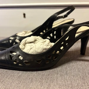 Svarta klackskor med utskurna detaljer - Snygga svarta klackskor från Ellos med spetsig tå och coola utskurna mönster på sidorna. Skorna har en smal rem runt hälen med spänne och är gjorda i syntetmaterial. Perfekta för dig som vill sticka ut med en elegant och modern look.