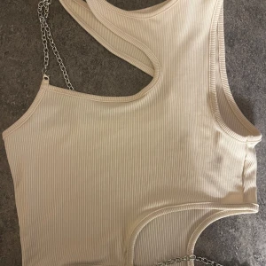 Beige ribbad linnetopp med kedjedetalj - Snygg beige/mörk vit ribbad linnetopp med silverfärgad kedja som axelband och runt midjan. Toppen är ärmlös och croppad.