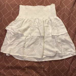 Vit volangkjol från 157 - Supersöt vit kortkjol från 157 med bred smock i midjan och dubbla volanger. Kjolen är luftig och har en flowy känsla, perfekt för varma dagar. Enkel att matcha med crop tops eller t-shirts för en chill look.