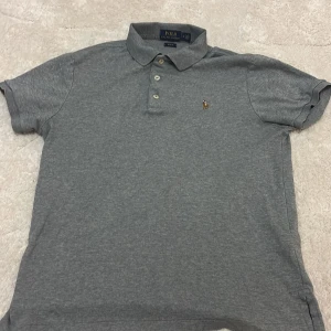 Grå pikétröja Polo Ralph Lauren M - Klassisk grå pikétröja från Polo Ralph Lauren i slim fit-modell. Tröjan har korta ärmar, krage med tre knappar och den ikoniska broderade polospelaren på bröstet. Tillverkad i mjuk bomull som är skön mot huden.