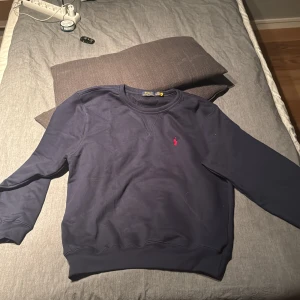 Mörkblå sweatshirt från Polo Ralph Lauren - Klassisk mörkblå sweatshirt från Polo Ralph Lauren i bomull. Tröjan har rund halsringning, långa ärmar och den ikoniska röda broderade loggan på bröstet. Perfekt för en chill och stilren look. Helt ny!