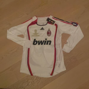 AC Milan långärmad Kaka' 22 tröja - AC Milan långärmad bortatröja från Adidas med Kaka' 22 på ryggen. Tröjan är vit med röda och svarta detaljer, Champions League-märke på ärmen och Bwin som sponsor på bröstet. Tillverkad i polyester, perfekt för dig som älskar fotboll och klassiska retrotröjor. Tröjan är också inge använd ännu.