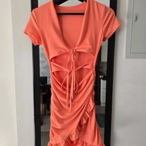 Koralldröm med snörning och volang - Superfin korallfärgad kortklänning med djup v-ringning, snörningsdetaljer framtill och volangkant nertill. Klänningen har korta ärmar och en snygg draperad effekt längs sidan. Perfekt för dig som vill sticka ut med en färgstark look!