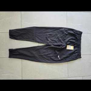 Svarta träningsbyxor från Nike - Svarta träningsbyxor från Nike med klassisk vit swoosh-logga på benet. Byxorna har resår i midjan och vid bensluten för en skön passform. Perfekta för chill eller träning, tillverkade i mjukt och lätt material.