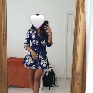 Blåvit kortklänning med knyt i midjan - Supersnygg blå kortklänning med stora vita mönster och knytband i midjan. Klänningen har skjortkrage, trekvartsärm och en lätt utställd kjol. Perfekt för dig som gillar en fräsch och trendig look med lite extra edge. Funkar för både xs & s