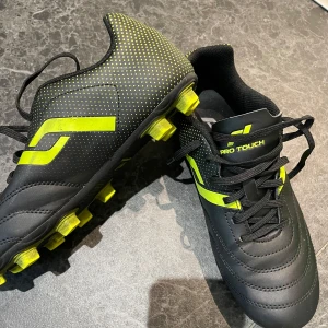 Pro Touch svarta fotbollsskor S - Svarta fotbollsskor från Pro Touch med neongula detaljer och snörning. Skorna har mönstrad ovandel med små gula prickar och robusta dobbar för bra grepp på planen. Tillverkade i syntetmaterial och gummi. Perfekta för dig som vill sticka ut på fotbollsplanen.