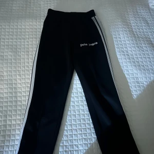 Palm Angels svarta mjukisbyxor med vita ränder - Svarta mjukisbyxor från Palm Angels med vita sidoremsor och logga tryckt på framsidan. Byxorna har elastisk midja, raka ben och dragkedja vid benslut. Snygg streetstil och sköna att bära, perfekta för chill eller träning.