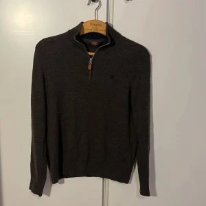 Mörkbrun merinoullströja Morris half zip - Snygg mörkbrun  tröja från Morris i 100% merinoull och i storlek XS-xxs Tröjan har half zip med läderdetalj vid dragkedjan och en liten broderad logga på bröstet. Långärmad och med ribbade muddar för en clean look. Hör av er vid frågor.