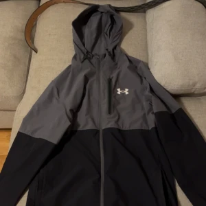 Under Armour svart/grå vindjacka S - Snygg svart och grå vindjacka från Under Armour med huva och hel dragkedja. Jackan har en bröstficka med dragkedja och logga på bröstet. Perfekt för blåsiga dagar och sportiga outfits.