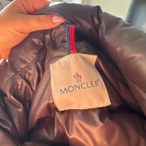 Grön Moncler pufferjacka - Säljer en brun pufferjacka från Moncler med glansig finish. Jackan har dragkedja och tryckknappar med Moncler-logga. Fodrad och riktigt varm, perfekt för kalla dagar. Klassisk design med hög krage och quiltade partier. Passar mig som vanligt vis är en 36/38 s/m