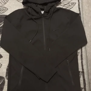 Svart vindjacka från Nike XL - Svart vindjacka från Nike i storlek XL med huva och dragsko. Jackan har hel dragkedja framtill, två sidofickor med dragkedja och diskret Nike-logga på bröstet. Perfekt för dig som gillar sportig och clean stil.