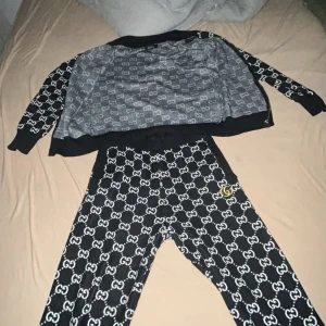 Svarta Gucci mjukisbyxor med logomönster - Svarta mjukisbyxor från Gucci med vitt GG-logomönster över hela byxan och en guldfärgad GG-detalj vid fickan. Byxorna har en avslappnad passform och resår i midjan. Perfekta för en trendig och bekväm streetstyle-look.