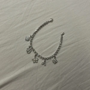 Glittrigt armband med berlocker - Ett silverfärgat armband med gnistrande stenar och fem olika berlocker, bland annat en blomma med pärla, löv och andra dekorativa former. Perfekt för dig som gillar detaljer och vill ha något som sticker ut.