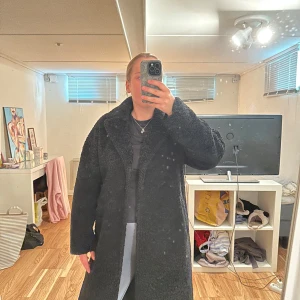 Svart lång teddyjacka med krage - Mysig svart teddyjacka i lång modell med bred krage och raka ärmar. Jackan har en fluffig och mjuk yta som ger en cool och avslappnad vibe. Perfekt för dig som vill ha en varm och trendig look under kyliga dagar. Finns fickor och knäpps med en knapp i midjan