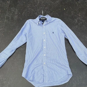 Blåvit randig skjorta Ralph Lauren XS - Klassisk blåvit randig skjorta från Ralph Lauren i slim fit. Skjortan har långa ärmar, knappar framtill och ikonisk broderad logga på bröstet. Tillverkad i stretchig bomull för extra komfort. Perfekt för en clean och stilren look. OBS den har några fläckar som visas i fotona 