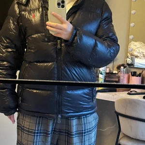 Svart dunjacka från Polo Ralph Lauren - Säljer en svart, glansig dunjacka från Polo Ralph Lauren med röd broderad logga på bröstet. Jackan har dragkedja framtill, flera fickor med dragkedja och en hög krage. Perfekt för kalla dagar och har en riktigt snygg, puffig look.