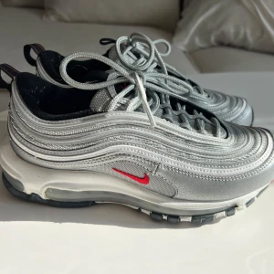 Nike Air Max 97 Silver Bullet - Säljer ett par ikoniska Nike Air Max 97 Silver Bullet med silvrig ovandel, röda detaljer och svart/vit sula. Skorna har den klassiska vågiga designen, synlig Air-enhet och snörning. 