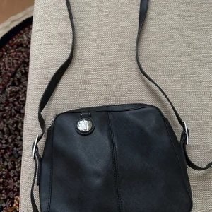 Svart axelväska från Armani Jeans - Snygg svart axelväska från Armani Jeans i stilren design. Väskan är tillverkad i skinnliknande material med silverfärgade metalldetaljer och dragkedja. Insidan har mönstrat foder med AJ-logga. Perfekt storlek för det viktigaste och justerbar axelrem.oanvänd helt ny.