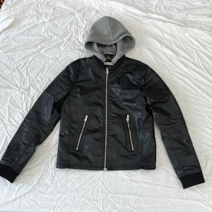 Svart jacka i skinnimitation  med grå huva H&M - Svart jacka i skinnimitation från H&M i storlek 152 med avtagbar grå huva. Jackan har två snedställda fickor med dragkedja, ribbade muddar vid ärmslut och nederkant samt coola sömdetaljer på axlarna. Perfekt för en streetinspirerad look.