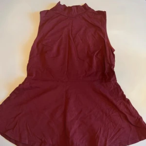 Vinröd ärmlös peplumtopp  - Vinröd ärmlös peplumtopp från Lager157 med hög hals och utställd kjol. Tillverkad i mjuk bomull med stretch för en bekväm passform. Enkel och stilren design som ger en snygg siluett.