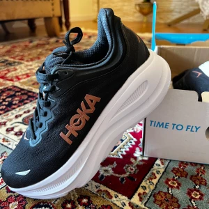 Hoka Bondi 9 svarta sneakers - Säljer ett par Hoka Bondi 9 sneakers i svart med vita sulor och detaljer i kopparfärg. Skorna har en chunky plattformssula, snörning och mesh-ovandel för extra andningsförmåga. Perfekta för dig som gillar stilrena och sportiga skor med maximal komfort. Köptes för 1800. Helt nya aldrig använda. Säljes då de var för små för mig