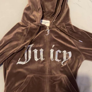Brun velour hoodie Juicy Couture XXS - Säljer en knappt använd,  brun hoodie från Juicy Couture i mjuk velour med glittrande stenar som bildar texten 'Juicy' på bröstet. Tröjan har huva, dragkedja med J-hänge och långa ärmar. Perfekt för dig som vill ha den ikoniska Y2K-looken.