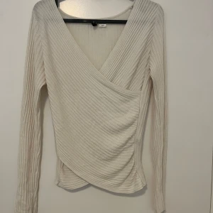 Ribbad omlottopp i beige - Snygg ribbad omlottopp i beige med lång ärm och djup v-ringning. Toppen har en figurnära passform och är tillverkad i mjukt material som känns skönt mot huden. Perfekt att styla med jeans eller kjol för en trendig look.