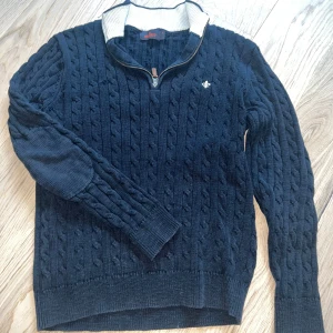 Mörkblå stickad tröja från Morris - Säljer en mörkblå stickad tröja från Morris med kabelstickat mönster, halv dragkedja och ribbade muddar. Tröjan har förstärkta armbågar och en vit detalj på insidan av kragen. Perfekt för kyliga dagar och fina middagar!