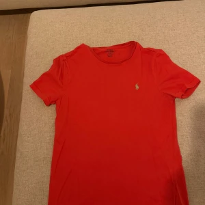 Röd t-shirt från Polo Ralph Lauren - Röd t-shirt från Polo Ralph Lauren med klassisk passform och korta ärmar. Ikoniska gröna loggan broderad på bröstet. Tillverkad i mjuk bomull som är skön mot huden. Perfekt för en avslappnad och stilren look.