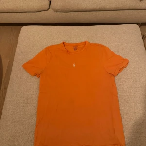 Orange t-shirt från Polo Ralph Lauren - Snygg orange t-shirt från Polo Ralph Lauren med klassisk passform och korta ärmar. Ikoniska broderade loggan i vitt på bröstet. Perfekt för dig som gillar färg och enkel stil.