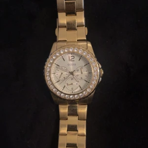 Guldig klocka med stenar från Guess - Snygg guldig klocka från Guess med länkarmband och glittrande stenar runt urtavlan. Urtavlan är silverfärgad med flera små visare och tydliga siffror. Perfekt accessoar för att lyfta din stil.