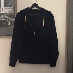 Ralph Lauren zip  - Marinblå hoodie från Polo Ralph Lauren med dragkedja, vit snörning i huvan och rött broderat logomärke på bröstet. Klassisk och stilren modell i mjuk bomull. Långärmad och med huva för extra komfort. 