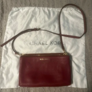 Röd axelväska från Michael Kors - Snygg röd axelväska från Michael Kors i äkta skinn med guldfärgade detaljer och dragkedja. Stilren design med justerbar axelrem och logga framtill. Perfekt storlek för det viktigaste och med ljus insida med diskret logomönster. Väskan har fått några små streck av mörk tuchpenna, som säkerligen går att ta bort, dock inget som stör intrycket av väskan eller funktionen, därav priset 🤗